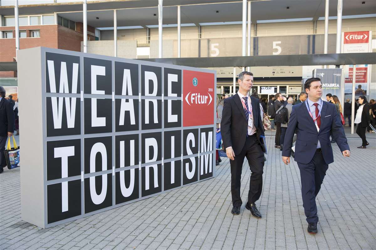 two men walk past We Are Tourism sign at FITUR