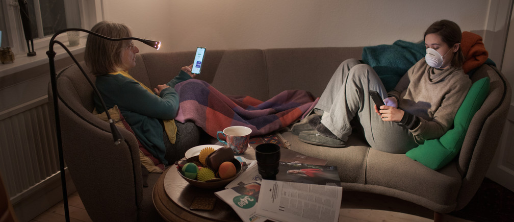 Two women with masks reading their smartphones in a livingroom